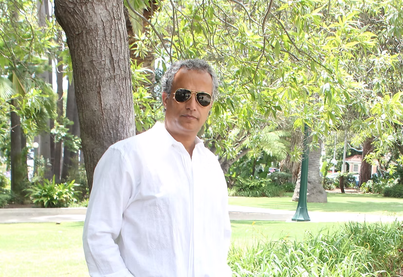 Man in white shirt and sunglasses standing outdoors near a tree.