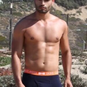 Shirtless man standing outdoors with mountains in the background.