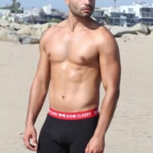 A shirtless man in black swim trunks standing on a sandy beach.